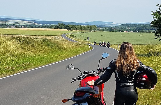 A person with long hair in bicycle leathers next to a motorbike watching other bikers in the distance along a long countryside road