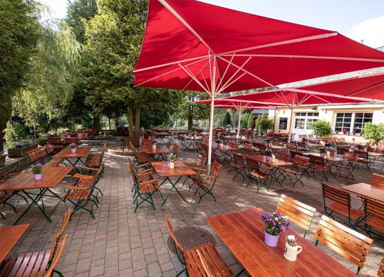 Großer Biergarten mit zahlreichen Holztischen und -bänken unter roten Sonnenschirmen, umgeben von alten Bäumen und blühenden Pflanzen. Die einladende Atmosphäre wird durch farbige Blumentöpfe auf den Tischen und das angrenzende Gebäude mit Fensterdekoration unterstrichen.