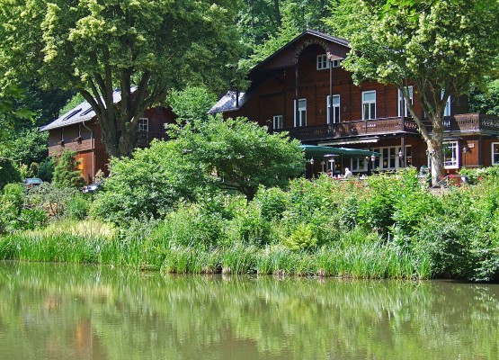 Ein hölzernes Ausflugslokal liegt idyllisch am Waldrand, umgeben von dichtem Grün und einem ruhig daliegenden Teich mit spiegelnder Wasseroberfläche. Die Terrasse des Hauses ist teils überdacht und von üppiger Vegetation umgeben.